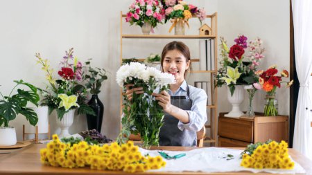 Foto de Florista femenina en delantal sosteniendo crisantemo blanco para crear y diseñar flores para arreglo ramo de flores en la mesa en su tienda de flores. - Imagen libre de derechos