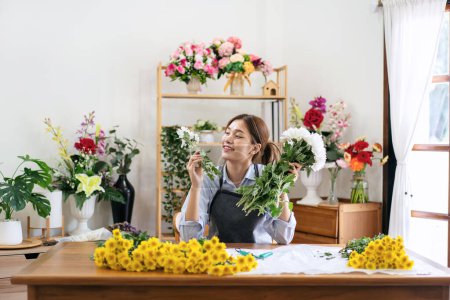 Téléchargez les photos : Fleuriste femelle dans tablier tenant chrysanthème blanc pour créer et concevoir floral pour arrangement bouquet de fleurs sur la table dans sa boutique de fleurs. - en image libre de droit