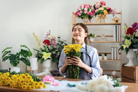 Photo for Female florist in apron holding yellow chrysanthemum and smell the flower with freshness while creating and designing floral to arrangement flower bouquet in her flower shop for delivery to customer. - Royalty Free Image