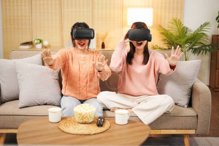 Foto de Mujer joven pareja lesbiana con suéter y gafas VR para jugar a videojuegos y ver la experiencia de la realidad virtual con disfruta mientras pasan tiempo juntos en la sala de estar en casa. - Imagen libre de derechos