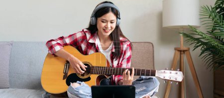 Photo for Young woman is playing guitar and practice to singing the song while wearing headphone to watching video tutorial on playing a musical instrument and sitting on comfortable the couch in living room. - Royalty Free Image