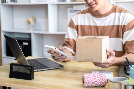 Photo for Male entrepreneur is using smartphone to checking client data and holding cardboard boxes for prepare to sending customer while working online shopping delivery at home office. - Royalty Free Image
