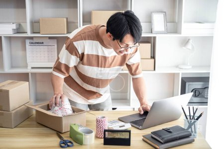 Photo for Male entrepreneur is using laptop to checking online orders and packing the product into cardboard boxes for prepare to sending customer while working online shopping delivery at home office. - Royalty Free Image