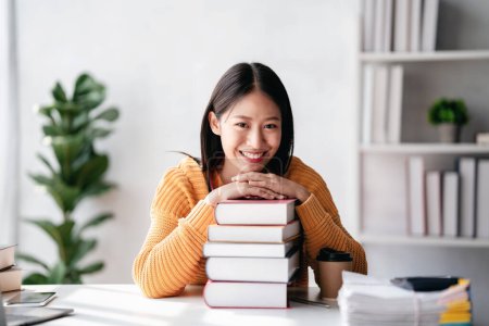 Téléchargez les photos : Concept d'enseignement à distance, Jeune femme portant un pull et souriant avec une pile de livres tout en lisant un livre et en étudiant une leçon d'éducation en ligne à la maison. - en image libre de droit