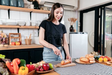 Téléchargez les photos : Femme tranchant le pain avec un couteau sur une planche à découper en bois et préparant des légumes frais avec des fruits sur la table pour cuisiner pour un petit déjeuner sain dans la cuisine moderne à la maison. - en image libre de droit