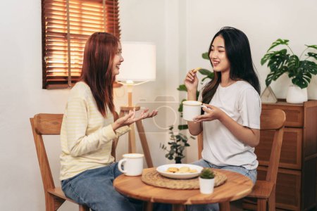 Foto de Mujer joven pareja lesbiana comiendo bocadillos y bebiendo chocolate caliente y hablando juntos mientras pasan tiempo para relajarse en la sala de estar en casa. - Imagen libre de derechos