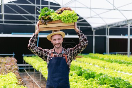 Foto de Agricultor de agronegocios y concepto de cultivo hidropónico, Hombre sonriendo y llevando canasta de verduras de ensalada en su cabeza después de cosechar el producto hidropónico en invernadero. - Imagen libre de derechos
