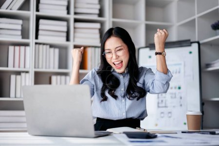 Accounting business concept, Accountant woman reading good news on laptop with excited face and raising fists to celebrate achievement while working about investment with business planning finance.