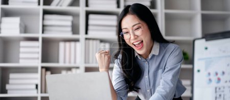 Accounting business concept, Accountant woman reads good news on laptop with excited and raising fist to celebrate achievement while working to typing about investment with business planning finance.