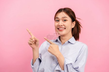 Téléchargez les photos : Jeune femme asiatique portant chemise à manches longues et utilisant les mains avec deux index pour pointer la publicité sur l'espace de copie isolé sur fond rose. - en image libre de droit