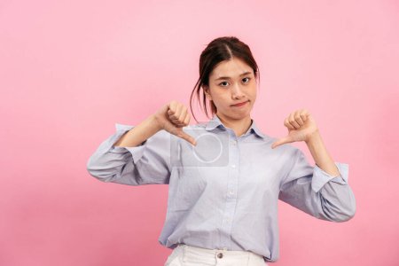 Photo for Young asian woman using hands and fingers while showing thumbs down for symbol of dislike or unlike with disappointed face isolated over pink background. - Royalty Free Image