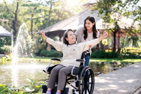 Senior im Rollstuhl und Tochter Familienkonzept, junge Asiatin schiebt Rollstuhl der Mutter zur Entspannung in den Garten, während Seniorin die Arme hebt, um frische Luft zu genießen und zu reden.