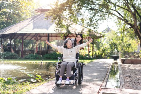 Foto de Senior en silla de ruedas y concepto de familia hija, Joven mujer asiática empujando silla de ruedas de la madre a la relajación en el jardín, mientras que la mujer mayor levantando los brazos para disfrutar del aire fresco y hablando. - Imagen libre de derechos