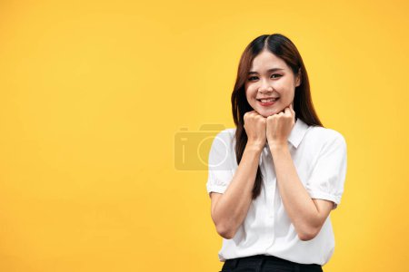 Téléchargez les photos : Jeune femme asiatique portant chemise blanche à manches courtes et toucher le menton avec les deux mains pour sourire tout en se tenant isolé sur fond jaune. - en image libre de droit