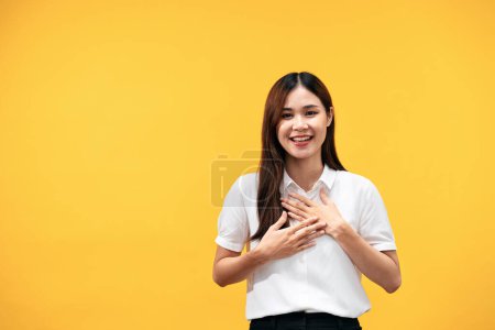 Foto de Joven mujer asiática con camisa blanca de manga corta y coloca ambas manos en su pecho mientras agradece el gesto y sonríe aislado sobre el fondo amarillo. - Imagen libre de derechos