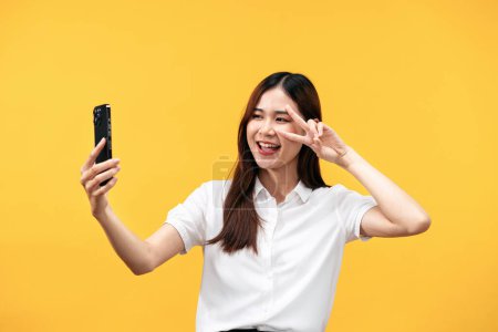 Téléchargez les photos : Jeune femme asiatique portant chemise blanche à manches courtes tout en utilisant un smartphone pour selfie avec signe v geste et sourire isolé sur fond jaune. - en image libre de droit