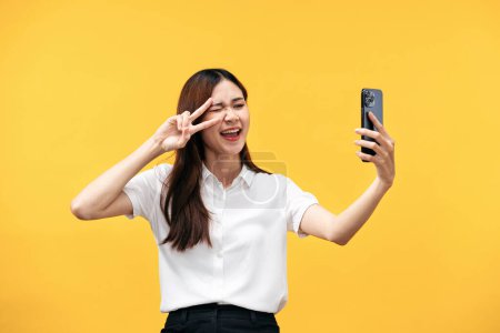 Foto de Joven mujer asiática que usa camisa blanca de manga corta mientras usa el teléfono inteligente para selfie con gesto de signo v y winkle con sonriente aislado sobre fondo amarillo. - Imagen libre de derechos