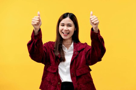 Foto de Joven mujer asiática con chaqueta roja y gran sonrisa después de tener buenas noticias para hacer pulgares hacia arriba gesto ambas manos y de pie aislado sobre fondo amarillo. - Imagen libre de derechos