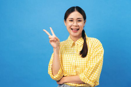 Foto de Joven mujer asiática con camisa amarilla de gingham con peinado trenzado y gafas para el brazo cruzadas mientras brilla y muestra v signo aislado sobre fondo azul claro. - Imagen libre de derechos