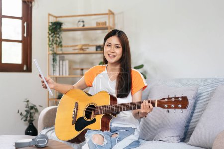 Photo for Young woman is holding a book of lyrics and chords to playing guitar and practice to singing the song while sitting on comfortable the couch in living room at home. - Royalty Free Image