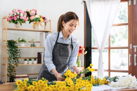 Téléchargez les photos : Fleuriste féminine dans tablier en sélectionnant chrysanthème jaune pour créer et concevoir floral pour arrangement bouquet de fleurs sur la table dans sa boutique de fleurs. - en image libre de droit