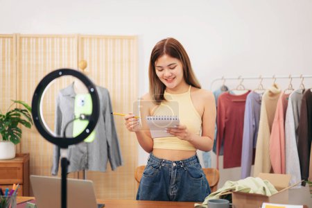 Photo for Woman designer entrepreneur showing clothes product on live online social media streaming and writing information of client in notebook while preparing product for shipping customer delivery at home. - Royalty Free Image