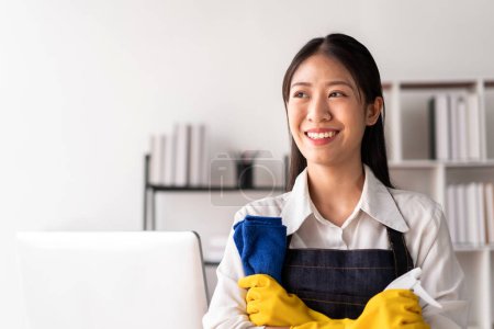 Téléchargez les photos : Femme au foyer dans le tablier portant des gants pour tenir bouteille de pulvérisation d'hygiène et tissu microfibre pour essuyer les meubles de nettoyage pendant le travail et le nettoyage dans la maison. - en image libre de droit