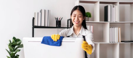 Téléchargez les photos : Femme au foyer dans le tablier portant des gants à l'aide de tissu en microfibre pour essuyer l'ordinateur et les meubles de nettoyage tout en travaillant et en nettoyant dans la maison. - en image libre de droit