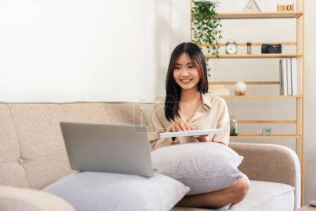 Photo for Female entrepreneur is sitting on big comfortable sofa and putting laptop and keyboard on pillow to typing business data while working in living room at home. - Royalty Free Image