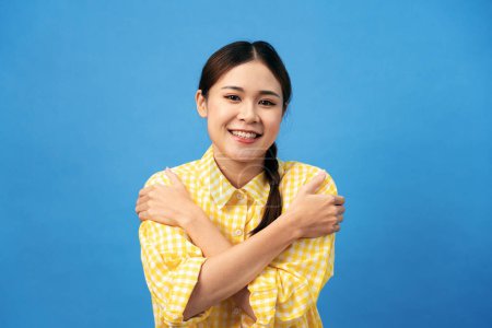 Photo for Young asian woman with braid hairstyle is beaming and embrace shoulders to hugging herself while standing isolated over light blue background. - Royalty Free Image