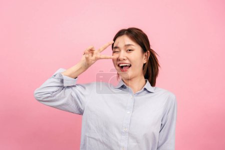 Foto de Joven mujer asiática con camisa de manga larga, mientras que el uso de la mano y los dedos para mostrar v gesto signo y winkle con sonriente aislado sobre fondo rosa. - Imagen libre de derechos