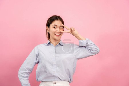 Foto de Joven mujer asiática con camisa de manga larga, mientras que el uso de la mano y los dedos para mostrar v gesto signo y winkle con sonriente aislado sobre fondo rosa. - Imagen libre de derechos