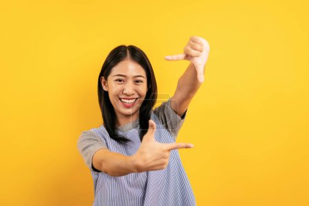 Téléchargez les photos : Jeune femme asiatique en vêtements décontractés souriant et faisant un geste de cadre photo pour prendre une photo sur l'appareil photo tout en posant comme un photographe isolé sur fond jaune. - en image libre de droit