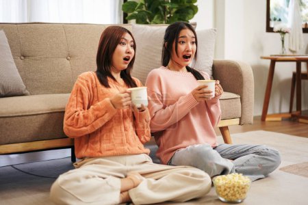 Photo for Young woman lesbian couple in sweater sitting on the floor to watching scary horror cinema together with shocked face and drinking hot chocolate with delicious popcorn while spending time together. - Royalty Free Image