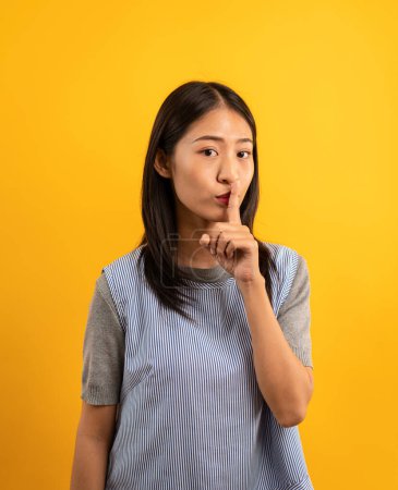 Foto de Joven mujer asiática en ropa casual haciendo un gesto silencioso para decir silencio para tranquilo y secreto con tocar el dedo en la boca aislado sobre fondo amarillo. - Imagen libre de derechos