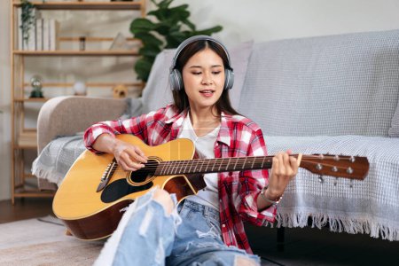 Téléchargez les photos : Jeune femme joue de la guitare et chante la chanson tout en portant un casque à écouter de la musique et assis sur le sol dans le salon à la maison. - en image libre de droit