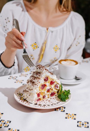 Close-up of a woman's hand taking a piece of fruit cream cake, with a cappuccino on the table