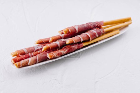 Elegante Grissini-Brotsticks in Prosciutto-Verpackung auf weißem Teller vor strukturiertem Hintergrund