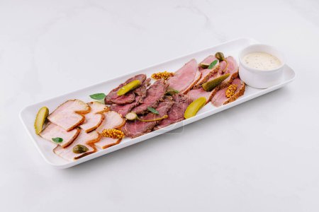 Photo for Gourmet platter of sliced deli meats served with pickles and a creamy dip on a white background - Royalty Free Image