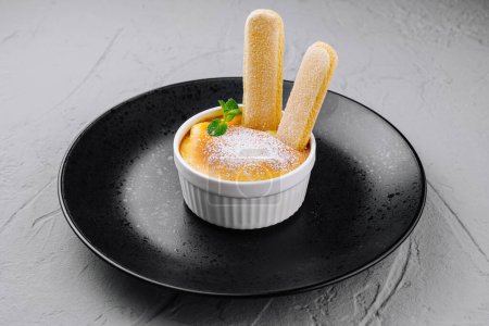 Classic creme brulee in a white ramekin with ladyfingers, dusted with powdered sugar on a dark plate