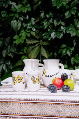 White ceramic jugs and cups with fruit on a table, green foliage background