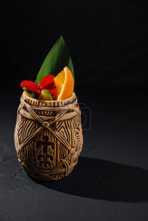 Taza tiki tradicional adornada con vibrante flora tropical y decoración de cítricos sobre fondo oscuro