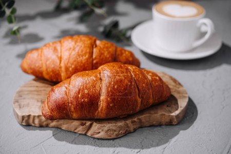 Two croissants on a wooden board paired with a cappuccino in a white cup, shot in daylight