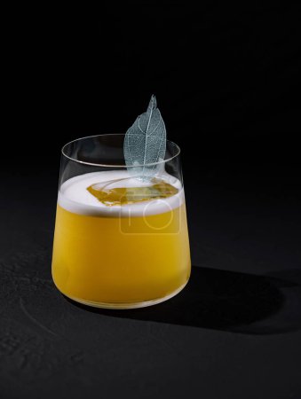 Modern cocktail featuring a frothy top and a delicate leaf garnish, presented on a dark backdrop