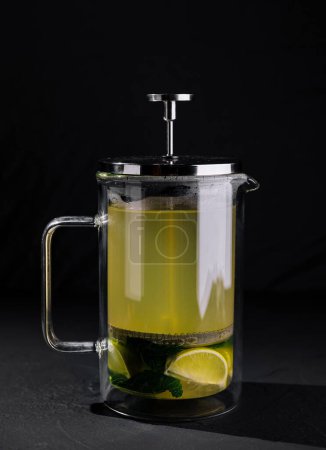 Clear glass french press filled with hot lime tea, mint leaves, and slices of lime on a dark background