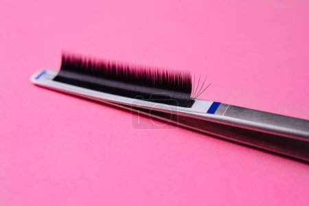 Detailed shot of individual fake eyelashes on tweezers, set against a vibrant pink backdrop
