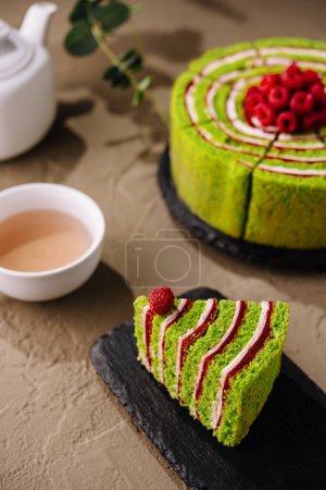 Tranche de gâteau de matcha au thé vert vibrant, décorée de framboises, à côté d'un gâteau complet et d'une tasse de thé