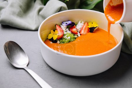 Bowl of creamy soup garnished with edible flowers and fresh strawberries on a sleek surface