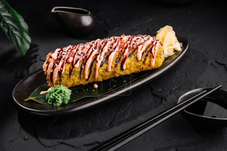 Delicious tempura sushi roll garnished with sauce, served with ginger and wasabi on a sleek black plate