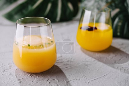 Zwei stiellose Gläser mit lebendigem Orangensaft und Eis, garniert mit Beeren, auf einer strukturierten Oberfläche